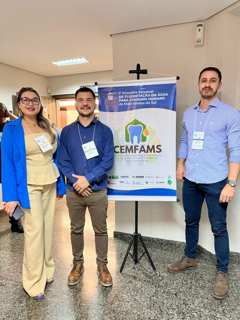 Coordenador da CATESA, Leandro Caldo (à direita), representante da AGEMS no Comitê Estadual de Monitoramento da Fluoretação da Água para o Consumo Humano posa ao lado de banner de encontro realizado em 2021 com mais dois integrantes.