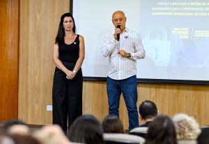 Presidente da AGEMS, Carlos Alberto de Assis, abre o 1º Fórum LGPD AGEMS, tendo ao lado a encarregada de dados, Analupe Urbieta. Em primeiro plano, com efeito desfocado, aparecem de costas algumas pessoas da plateia.