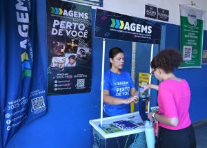 Atendente da Ouvidoria Katia Oliveira conversa com uma mulher jovem, atrás de um totem do projeto AGEMS Perto de Você. Ela usa uniforme do projeto e a jovem usa camisa rosa, calça preta e óculos de aros vermelhos.