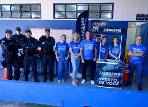 Policiais Militares e atendentes da Ouvidoria da AGEMS posam no corredor de uma escola municipal, junto a banner e totem do projeto AGEMS Perto de Você.