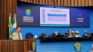 Servidor da AGEMS Paulo fala, da tribuna da Assembleia, olhando para a plateia. Ao fundo aparece a Mesa Diretora com autoridades e, atrás delas, slide com dados técnicos da AGEMS em relação a fiscalização de energia elétrica.