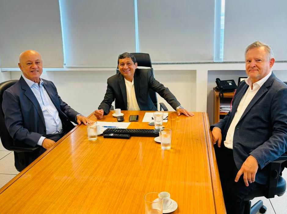Três homens reunidos em uma mesa retangular, de madeira, em sala com parede e cortina claras. Usam ternos escuros e camisas claras e sorriem posando para foto. São os diretores da AGEMS, Carlos Alberto de Assis e Matias Gonsales, e o diretor-geral da Agência Nacional de Mineração, Mauro Henrique Moreira Sousa.