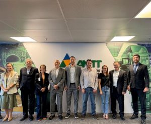Diretores da AGEMS Carlos Alberto, Matias Gonsales, Iara Marchioretto e Caroline Tomanquevez posam para foto com dirigentes da ANTT, diante de um painel que toma toda a parede, com a logomarca da ANTT sobre imagens em tom verde de rodovia e ônibus. Os homens vestem ternos e as mulheres roupa social.