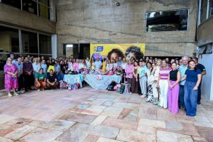 presidente da AGEMS, parceiras do projeto Lenços de Luz e servidores posam atrás de uma mesa com lenços e demais acessórios coloridos de cabeça.