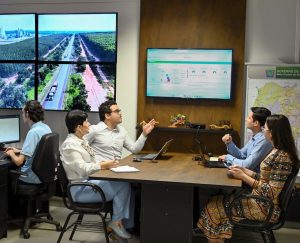 Dois homens e duas mulheres integrantes da Regulação Econômica conversam em torno de uma mesa. Utilizam notebooks, tablets e agendas, observam numa TV um painel de dados com números e gráficos. Ao fundo, um funcionário manuseia um computador, tendo ao alto uma painel formado por grandes TVs onde se vê a imagem da BR-262, com floresta de eucalipto ao lado e uma indústria ao longe.