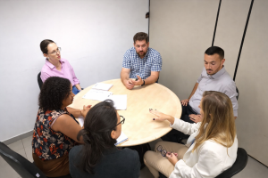 Ao redor de uma pequena mesa redonda, a diretora da AGEMS Iara, e o prefeito de Pedro Gomes, Murilo Jorge, e suas equipes conversam sobre a gestão de resíduos sólidos no município. Ao todo são seis pessoas, sendo quatro mulheres e dois homens.