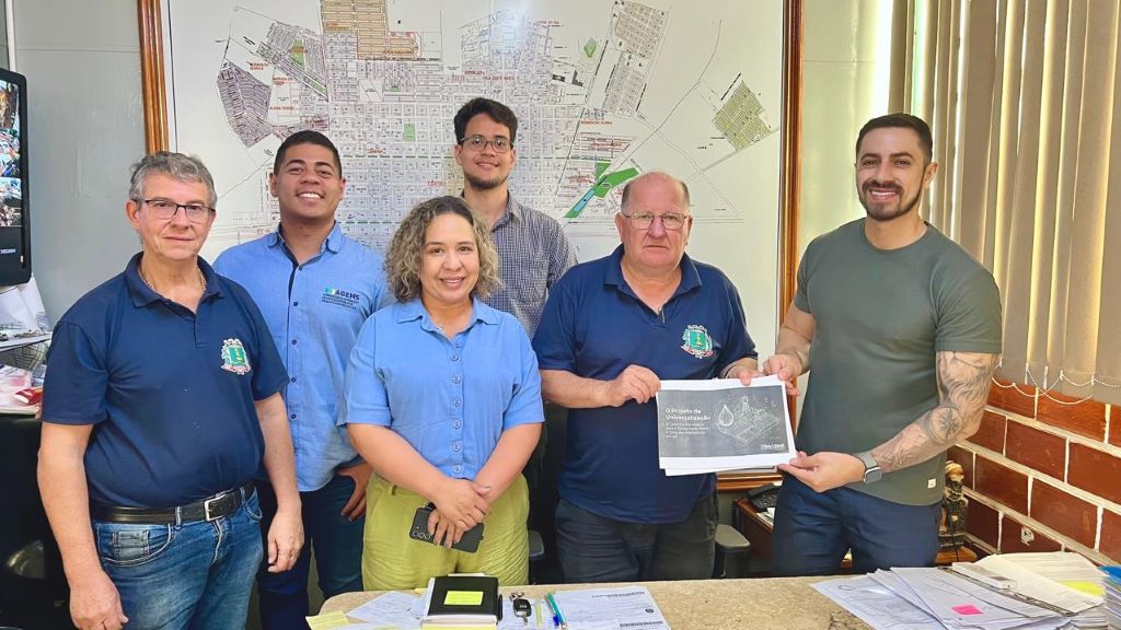 Equipe da AGEMS posa com secretário de infraestrutura de Sidrolândia segurando projeto sobre soluções alternativas de saneamento, no gabinete do secretário.