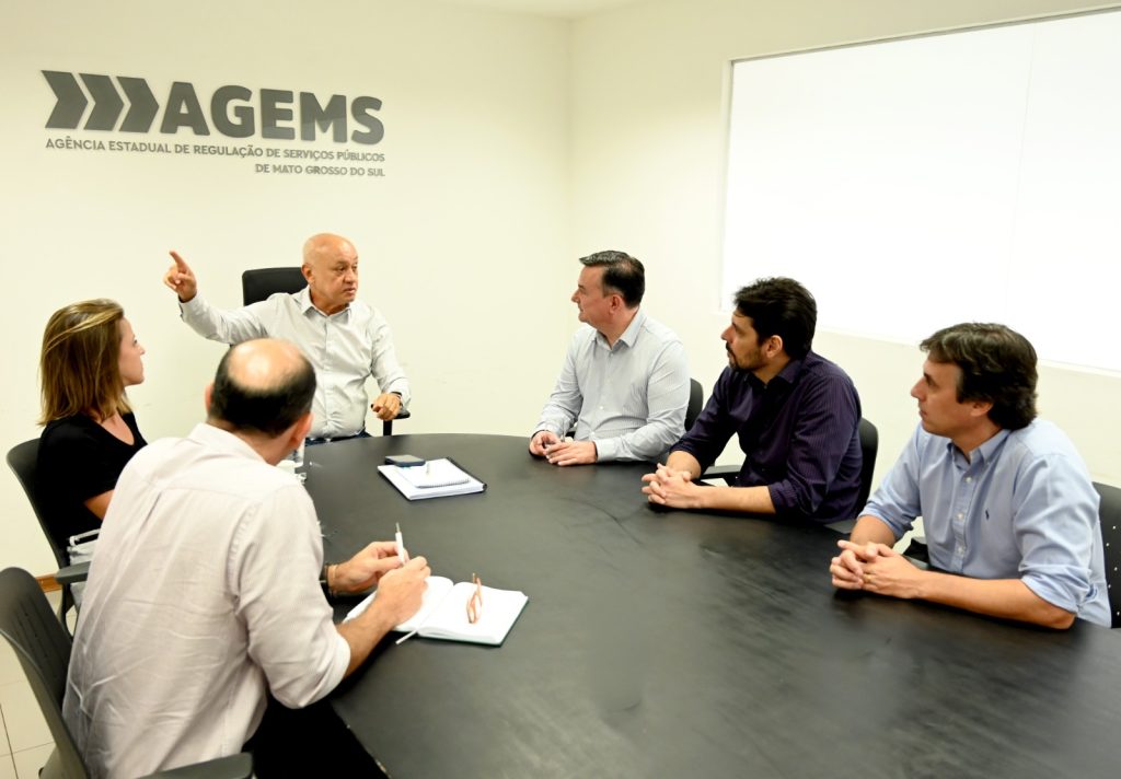Cinco homens e uma mulher usando roupas claras e escuras sentados em uma mesa de reunião preta.