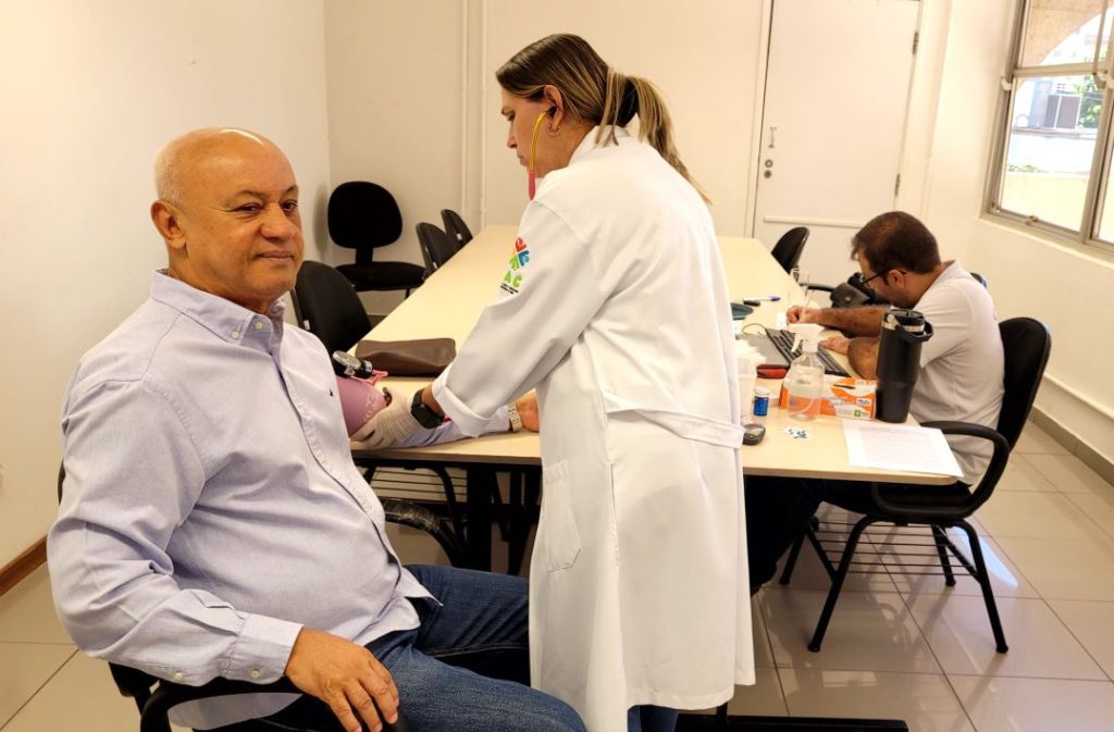 Carlos Alberto de Assis, sentado, usando camisa social e jeans, tem a pressão aferida por profissional usando jaleco branco, enquanto outro profissional faz anotações na mesa ao lado.