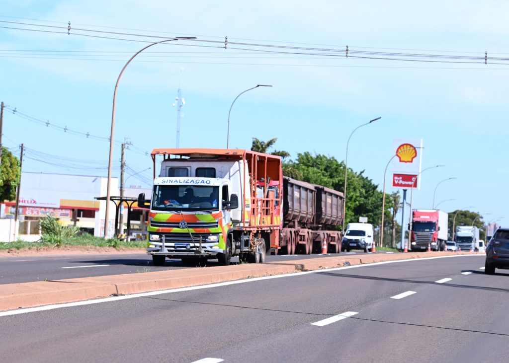 Uma estrada sendo sinalizada, com um caminhao branco e laranja com uma placa de posto de combustivel ao fundo.