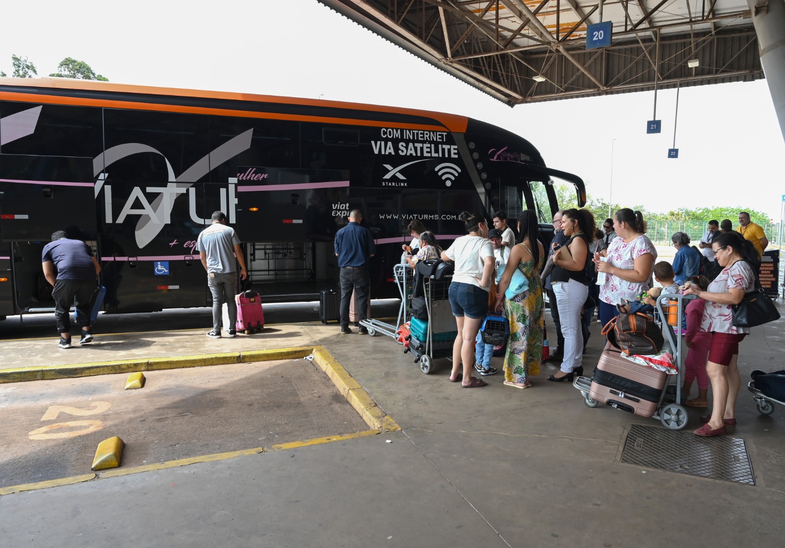 Na plataforma da rodoviária de Campo Grande, uma fila de passageiros despacha bagagem e se prepara para embarcar em ônibus da Viatur, de cor preta, com detalhes alaranjados e rosa.