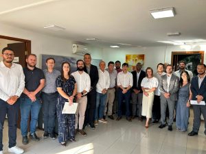 Na antessala do gabinete de direção da AGEMS, o presidente Carlos Alberto, outros servidores e representantes da Arauco e concessionária Way 112 posam para foto. O grupo é formado por 15 homens e 3 mulheres.